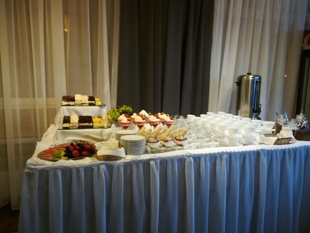 Na zdjęciu nakryty białym obrusem duży stół, na którym stoją przygotowane filiżanki, talerzyki oraz ciasto.