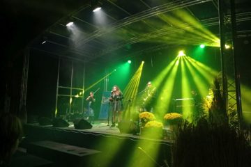 Zdjęcie z występu. Scena z zielonym oświetleniem, na scenie stwoi wokalista i dwóch gitarzystów