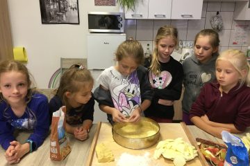 sześć dziewczynek przy stole, na stole artykuły spożywcze, stolnica, a na niej forma do pieczenia ciasta, dzieci robią szarlotkę