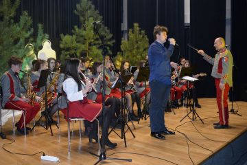 zdjęcie Młodzieżowej Orkiestry Dętej z Poniatowej na scenie obok dyrygent oraz solista w tle choinki