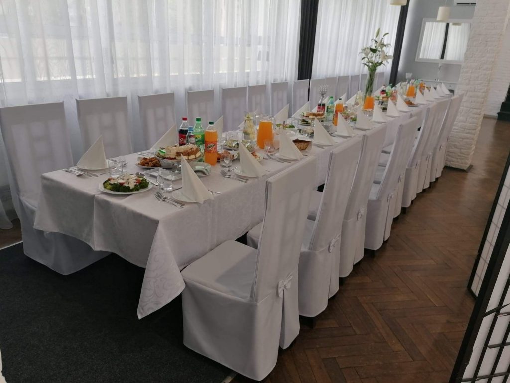 Na zdjęciu wnętrze kawiarni Klubowa, zastawiony potrawami stół, dookoła stoją białe krzesła.