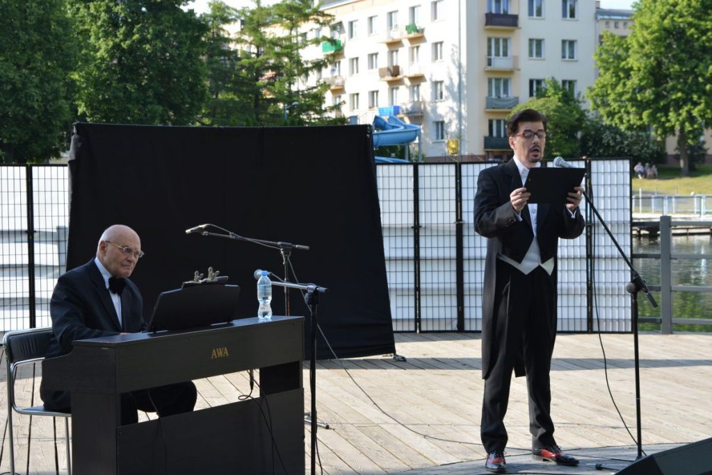 zdjęcie dwaj mężczyźni w garniturach jeden gra na pianinie drugi śpiewa do mikrofonu w tle budynki