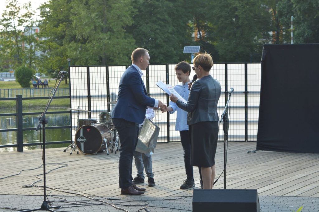 zdjęcie mężczyzna w marynarce wręczający nagrodę na scenie chłopcu obok kobieta z mikrofonem