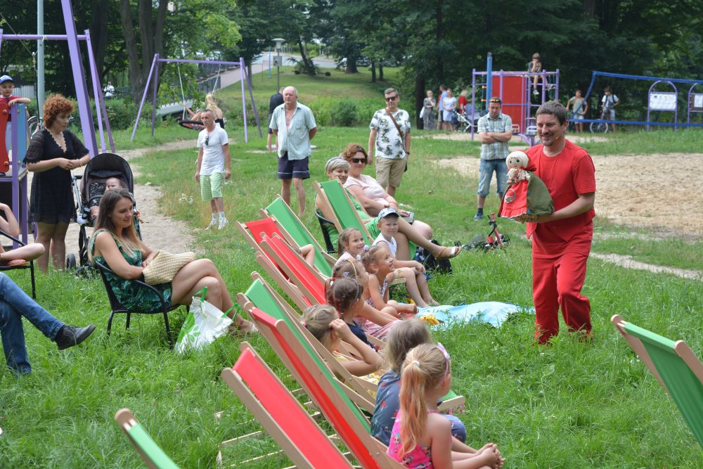 zdjęcie aktor w czerwonym stroju z lalką w ręce stoi obok dzieci siedzących na leżakach na trawie