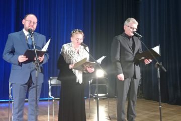 Trzy osoby (dwóch mężczyzn i kobieta) stoją oświetleni na scenie i trzymają w rękach podkładki z których czytają wiersze. Za nimi w tle niebieskie zasłony