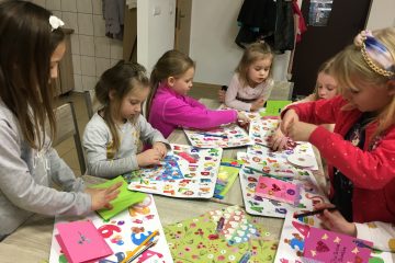 6 dziewczycnek siedzi przy stole i dekoruje kolorowymi naklejkami swoje laurki