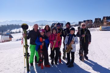 Uczestnicy Orkiestry Dętej na stoku w górach, ubrani w stroje narciarskie, w tle góry niebo i śnieg
