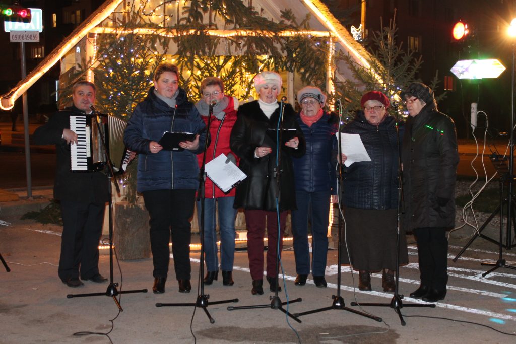 szopka bożonarodzeniowa a w niej drzewka, przed szopką stoją kobiety, śpiewają do mikrofonów