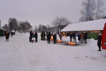 Ludzie stojący przy ognisku, na ziemi dużo śniegu, w tle osoby odwiedzjące kiermasz