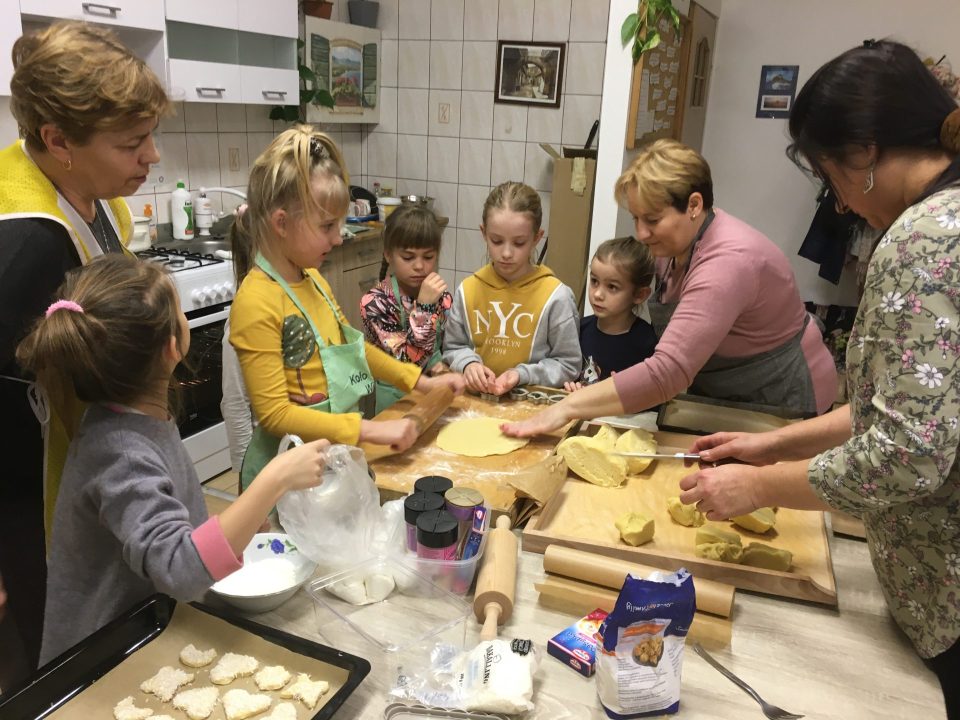 zdjęcie przedstawia grupę dzieci, które robią ciastka, na stole stoi mąką, stolnica, wałek do ciasta, naczynia, dzieciom pomaga opiekunka