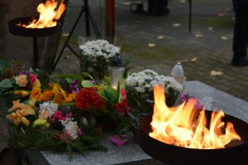 zdjęcie przedstawia kwiaty leżące na pomniku oraz płonące znicze
