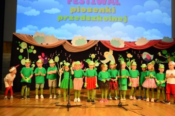 grupa dzieci na scenie, dzieci ubrane są w zielone kostiumy żab, w tle znajduje się kolorowa dekoracja