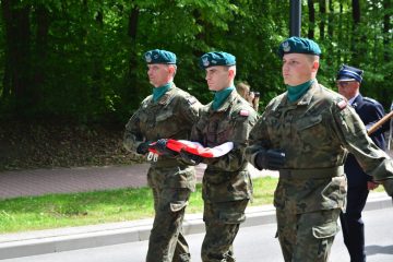 trzech maszerujących żołnierzy w mundurach, jeden z nich trzyma w rękach flagę Polski