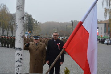 Dwóch mężczyzn stojących przed pomnikiem. Jeden z nich jest ubrany w mundur wojskowy, na głowie ma czapkę. Dłonią oddaje hołd. Drugi mężczyzna w czarnym płaszczu pochyla głowę do przodu. Na pierwszym planie zdjęcia widzimy flagę Polski.